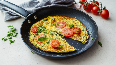 Eine angeschnittene Zucchinipizza in einer Pfanne. Links neben der Pfanne liegen einige Kräuter. Über der Pfanne ein Tuch und daneben weitere Tomaten.