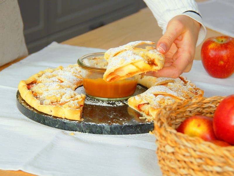 Eine ringförmige Apfeltarte mit Lotus-Creme, mit einer Schüssel Karamell-Dip in der Mitte. Der Ring ist angeschnitten und es ist eine Hand zu sehen, die sich ein Stück der Apfeltarte mit Lotus-Creme nimmt. Die Tarte ist mit Puderzucker bestäubt. Das runde Blech mit der Tarte steht auf einer Holzarbeitsfläche, die mit einem weißen Tuch bedeckt ist. Außerdem ist ein Korb mit Äpfeln zu sehen.