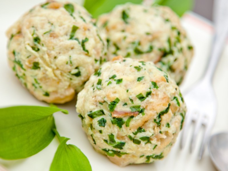 Drei Bärlauchknödel auf einem Teller. Daneben liegen Besteck und frischer Bärlauch.