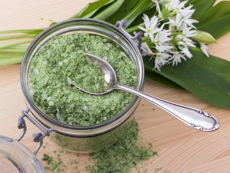 Ein Glas Bärlauchsalz mit einem Löffel, daneben Bärlauchblätter und Blüten. Nahaufnahme.