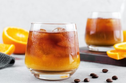 Zwei Gläser Bumble Coffee mit Eiswürfeln. Daneben liegen Orangen und Kaffeebohnen.