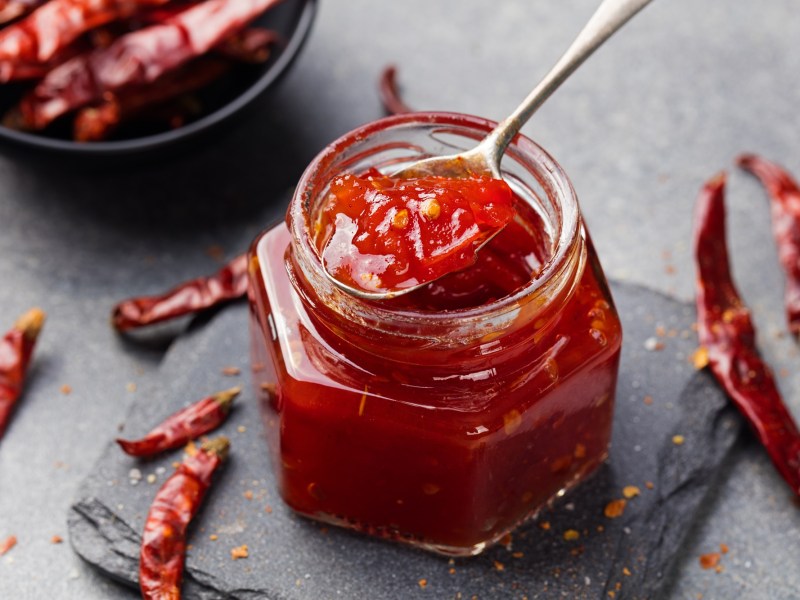 Ein Glas Chilimarmelade. Drum herum liegen Chilischoten.