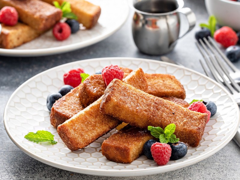 French-Toast-Stangen auf zwei Tellern, garniert mit Beeren, daneben Besteck.