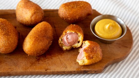 Mehrere Mini Corn Dogs aus dem Airfryer auf einem Holzbrett. Daneben steht ein Schälchen mit gelbem Dip.