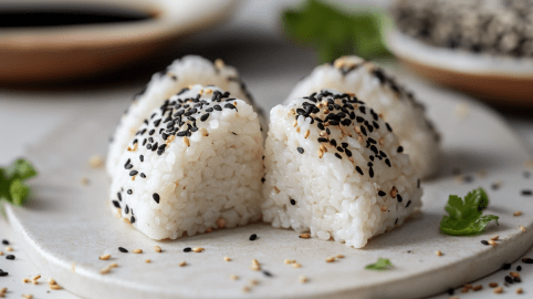 Onigiri ohne Noriblätter auf einer weißen Platte. Sie sind mit schwarzen und weißen Sesamkörnern bestreut. Neben ihnen liegen einige Kräuter, im Hintergrund sieht man eine große Schale mit Sojasoße.