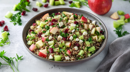 Ein großer Teller mit Quinoa-Apfel-Salat mit Sellerie und Cranberrys. Außerdem enthält der Salat frische gehackte Petersilie und Feta. Um den Teller herum liegen einzelne Cranberrys, Apfel- und Selleriestücke verteilt, sowie Petersilie. Im Hintergrund ist zudem ein ganzer Apfel erkennbar.