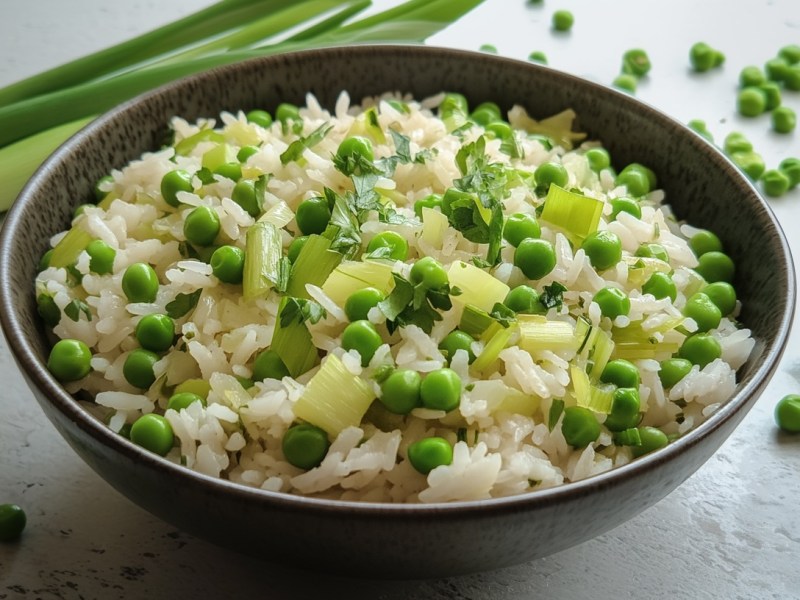 Eine Schüssel Reis mit Lauch und Erbsen, drumherum einige Erbsen und Lauchstangen.