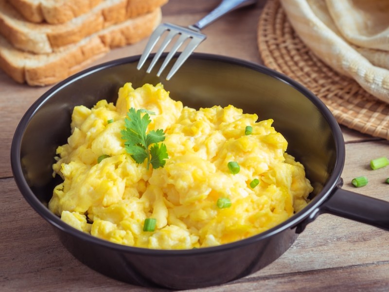 Rührei mit Hüttenkäse und Schnittlauch in einer kleinen Pfanne, daneben eine Gabel und Toastbrotscheiben.