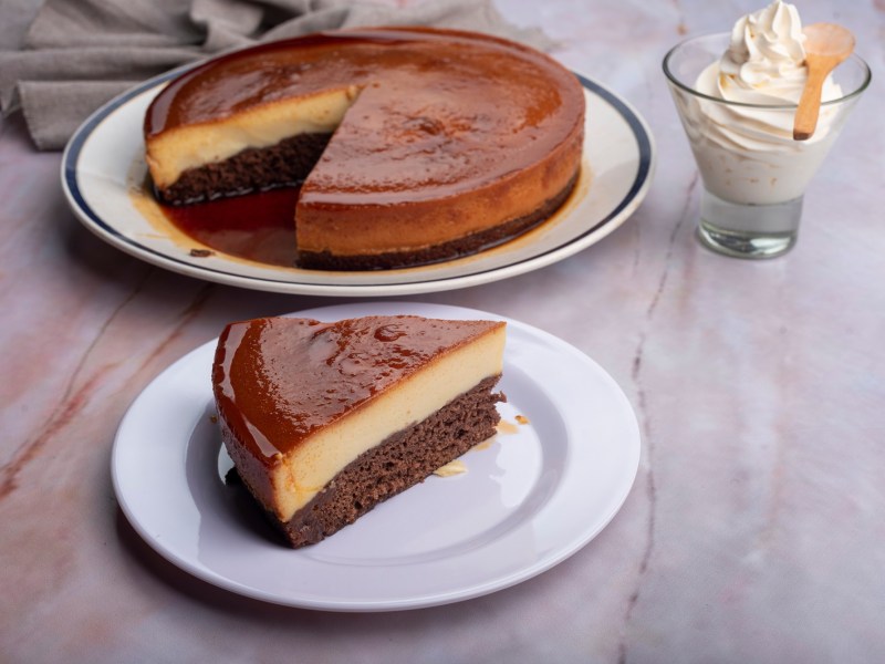 Ein Stück Schoko-Flan-Kuchen auf einem Teller. Im Hintergrund steht der restliche Kuchen.