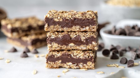 3 Schoko-Haferflocken-Riegel übereinander gestapelt, im Hintergrund einige Zutaten wie Schokolade und Haferflocken unscharf zu erkennen.