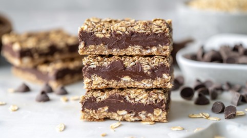 3 Schoko-Haferflocken-Riegel übereinander gestapelt, im Hintergrund einige Zutaten wie Schokolade und Haferflocken unscharf zu erkennen.