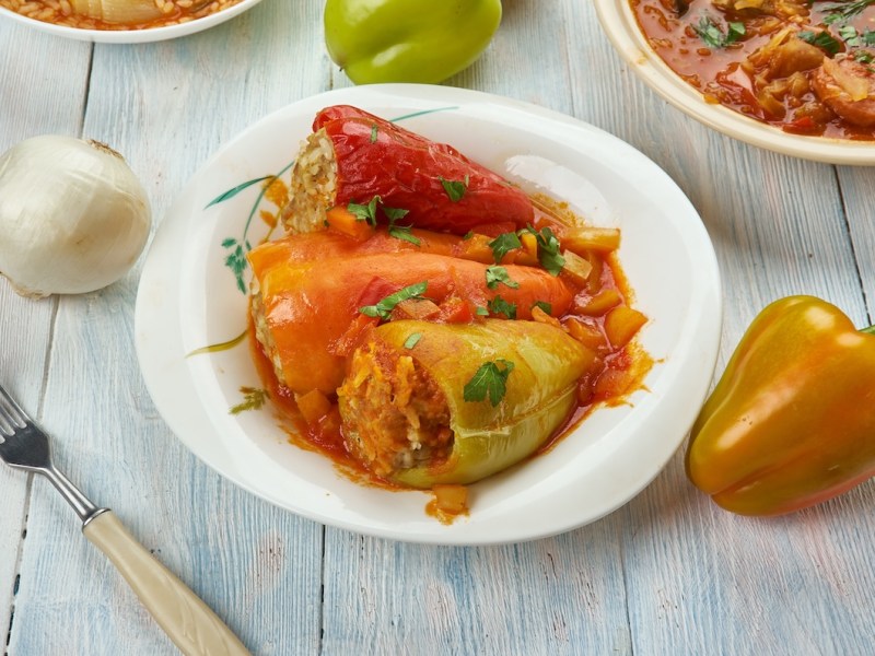 3 ungarische gefüllte Paprika mit Tomatensoße auf einem Teller, daneben eine Gabel, eine Paprika und eine Zwiebel sowie ein Topf mit Tomatensoße.