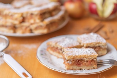 Ein paar Stücke ungarischer Apfelkuchen auf einem Kuchenteller, im Hintergrund weitere Stücke und Äpfel.