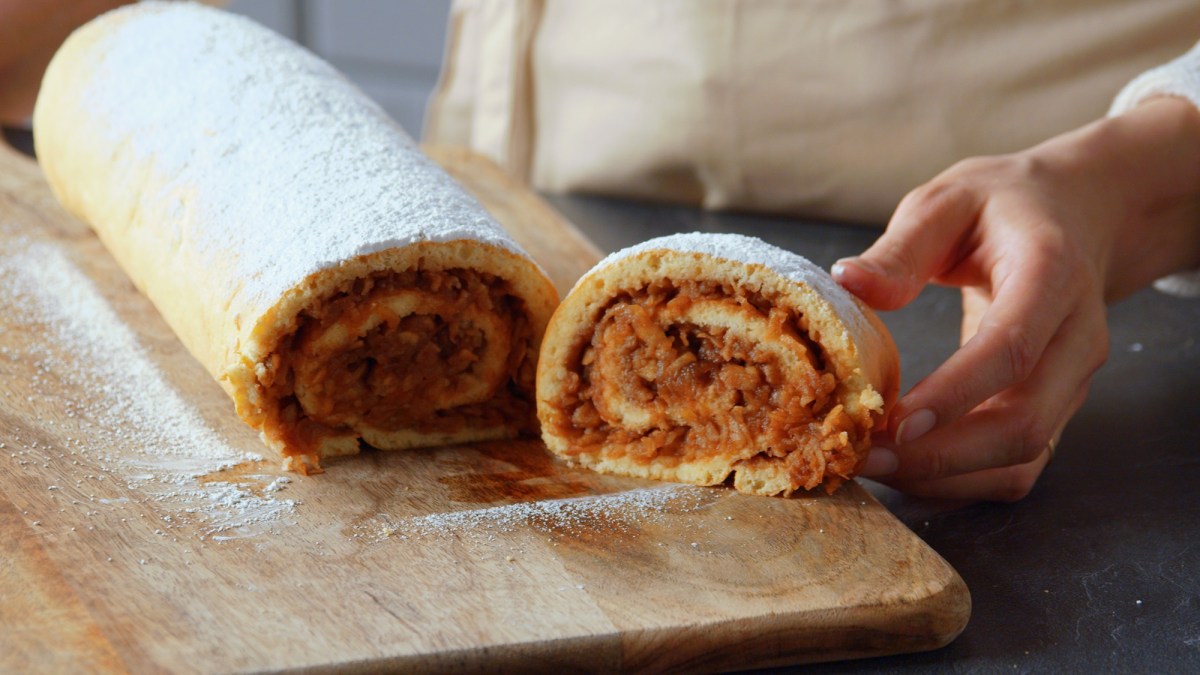 Ein Holzbrett, auf dem eine süße Apfelrolle liegt, die mit Puderzucker bestäubt ist. Man kann eine Hand erkennen, die ein abgeschnittenes Stück präsentiert.