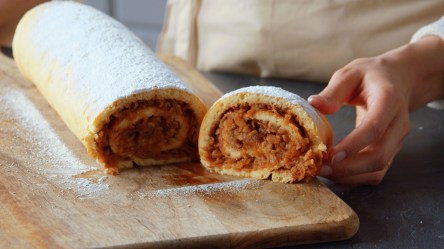 Ein Holzbrett, auf dem eine süße Apfelrolle liegt, die mit Puderzucker bestäubt ist. Man kann eine Hand erkennen, die ein abgeschnittenes Stück präsentiert.