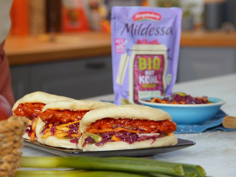 Bao Buns mit Rotkohl-Mangosalat und Hähnchen auf einem Teller. Im Hintergrund ist eine Tüte MIldessa Bio Rotkohl gekocht zu sehen.