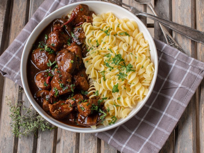 Ein Teller Brauhausgulasch mit Nudeln in der Draufsicht, der Teller steht auf einer karierten Serviette, daneben liegt Besteck.