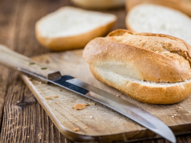 Ein aufgeschnittenes Brötchen, das im Airfryer aufgebacken wurde, auf einem Brett. Daneben ein Messer und im Hintergrund weitere Brötchen.