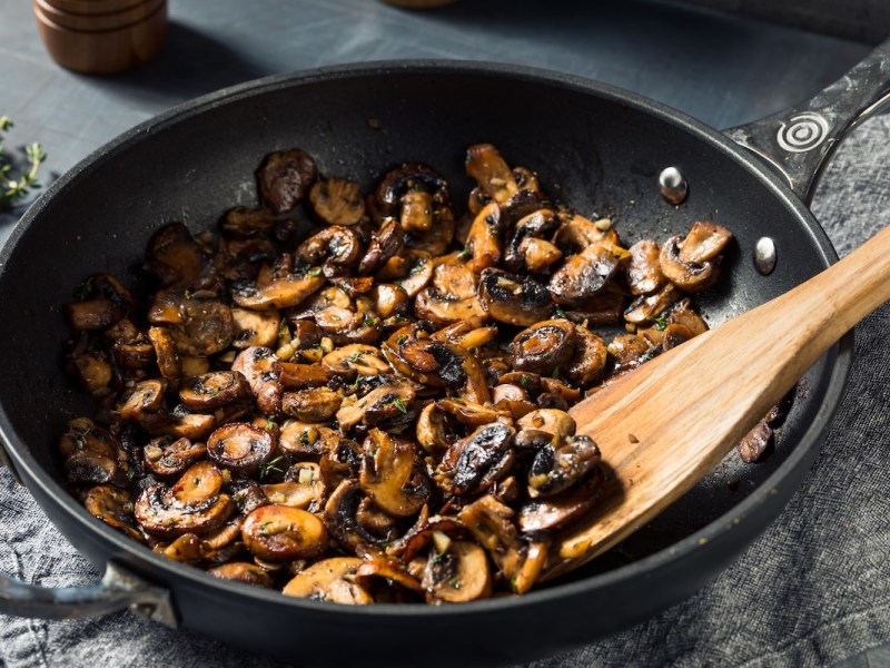 Champignons mit Kräuter-Chili-Soße in einer Pfanne mit einem Holzlöffel. Nahaufnahme.