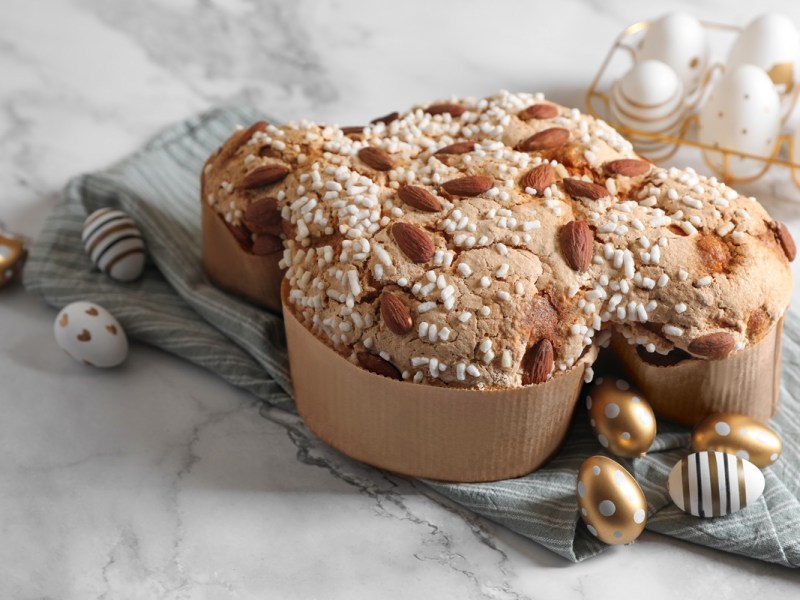 Colomba Pasquale: italienischer Osterkuchen in Taubenform in einer Papierhülle. Drum herum liegen dekorative Ostereier. Der Kuchen steht auf einem Leinentuch, das wiederum auf einer Marmorplatte drapiert ist.