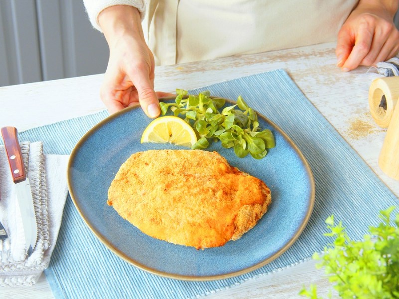 Ein Cordon Bleu mit Salami auf einem blauen Teller. Auf dem Teller liegt eine Zitronenspalte und etwas Feldsalat. Neben dem Teller liegt etwas Besteck und eine Salz und Pfeffermühle.
