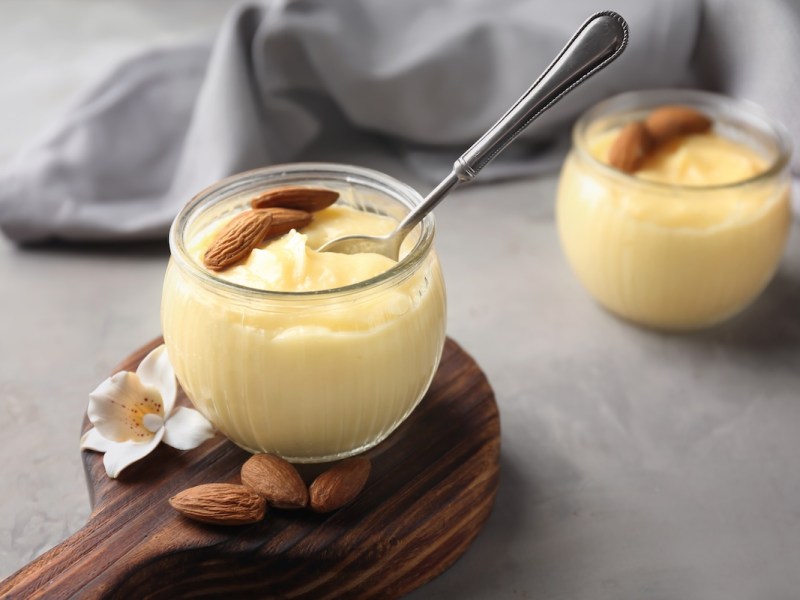 2 Gläser Eierlikör-Creme garniert mit Mandeln, in einer steckt ein Löffel. Dahinter eine Stoffserviette.