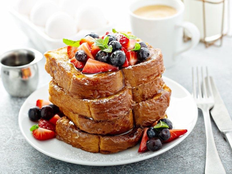 Ein Stapel French Toasts mit Eierlikör, garniert mit frischen Beeren, auf einem Teller. Daneben Besteckt, eine Kanne Sirup und eine Tasse.