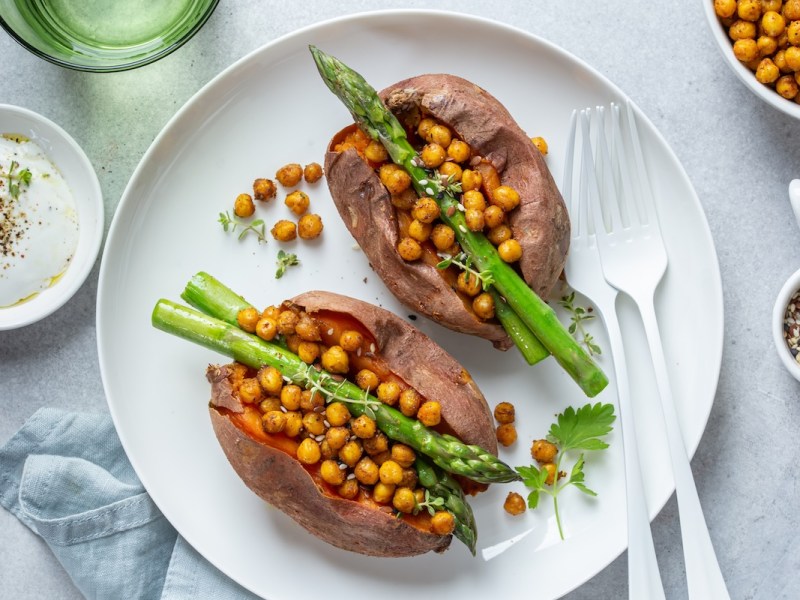 1 halbierte gebackene Süßkartoffel mit Spargel und Kichererbsen auf einem Teller, daneben weißes Besteck und Schüsseln mit Zutaten. Draufsicht.