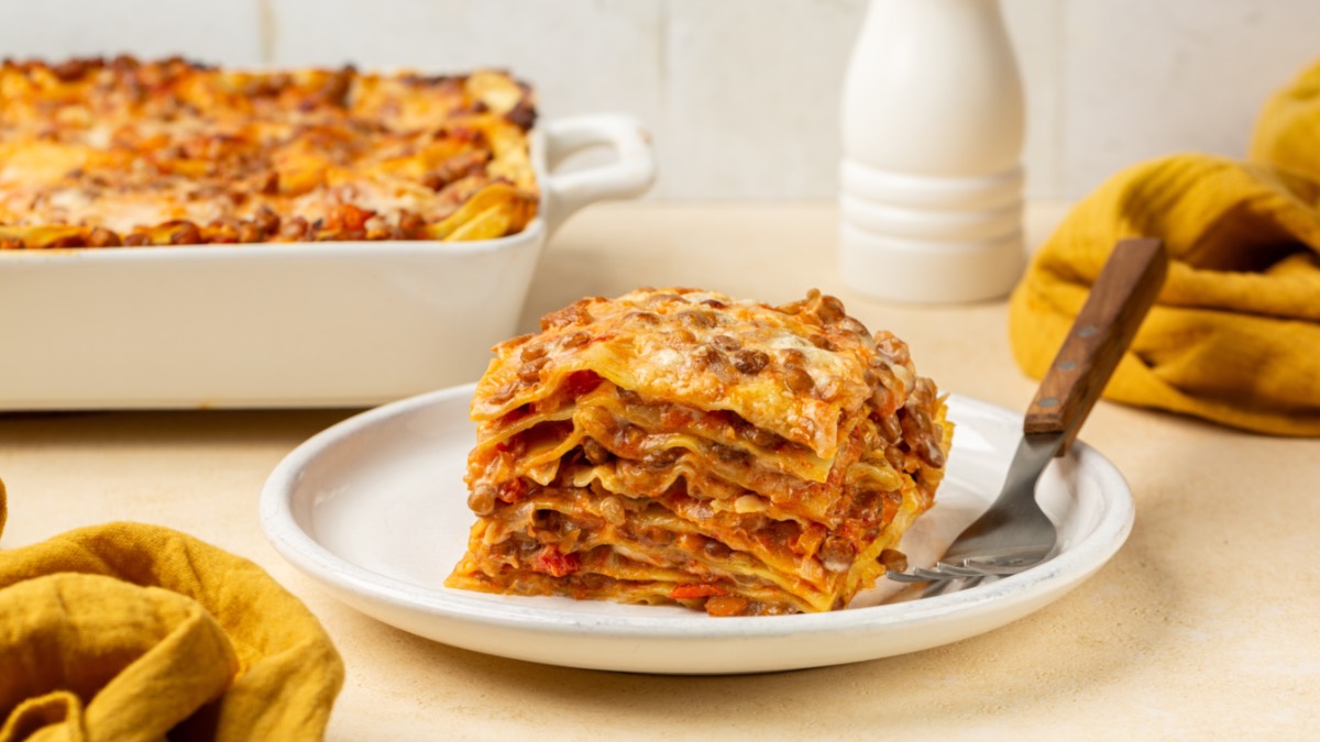 Ein Stück Linsen-Lasagne auf einem Teller. Im Hintergrund ist der Rest in einer Auflaufform zu sehen.