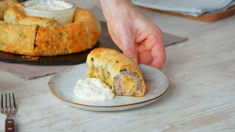 Ein Stück Weisskraut-Kartoffel-Strudel mit einer Portion Sour Cream auf einem Teller. Im Hintergrund kann man den restlichen, schneckenförmigen Strudel erkennen. In der Mitte des Strudels ist eine Schüssel mit Sour Cream platziert.