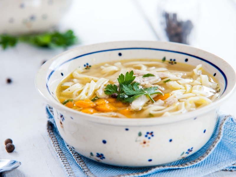 Eine Schüssel Nudelsuppe mit Gemüse auf einer Stoffserviette, daneben ein Löffel.