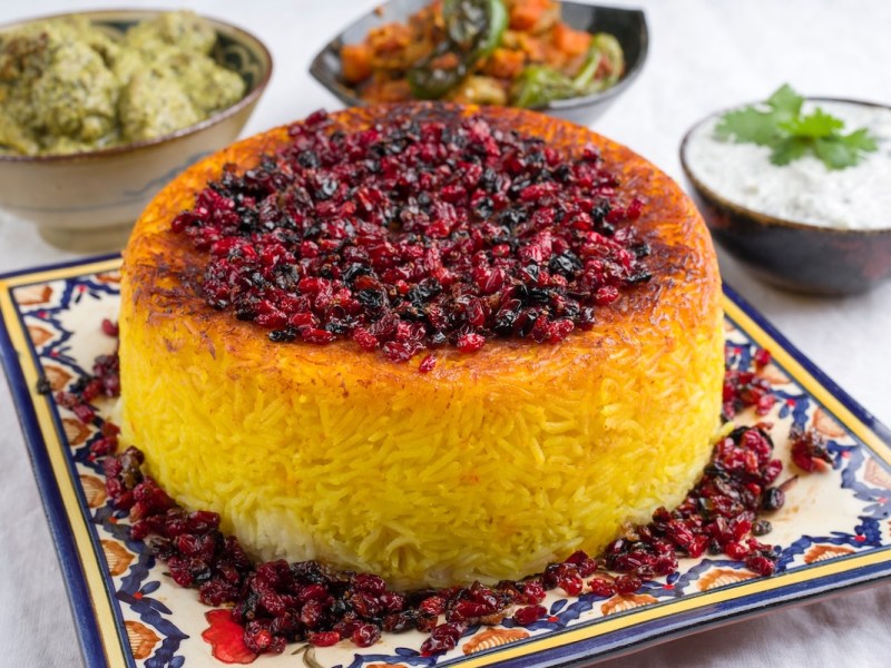 Persischer Reis Tahdig auf einem gemusterten Teller, garniert mit Trockenfrüchten, dahinter Schüsseln mit kleinen Speisen.