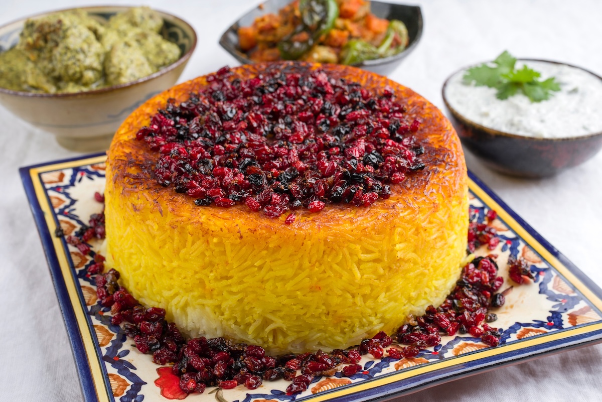 Persischer Reis Tahdig auf einem gemusterten Teller, garniert mit Trockenfrüchten, dahinter Schüsseln mit kleinen Speisen.