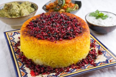 Persischer Reis Tahdig auf einem gemusterten Teller, garniert mit Trockenfrüchten, dahinter Schüsseln mit kleinen Speisen.