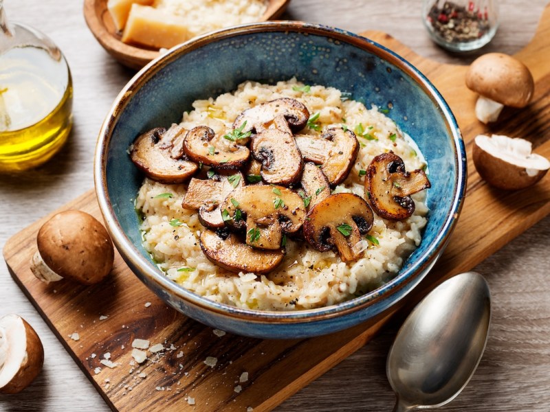 Eine Schüssel Pilzrisotto auf einem Brett, daneben Champignons und ein Löffel.
