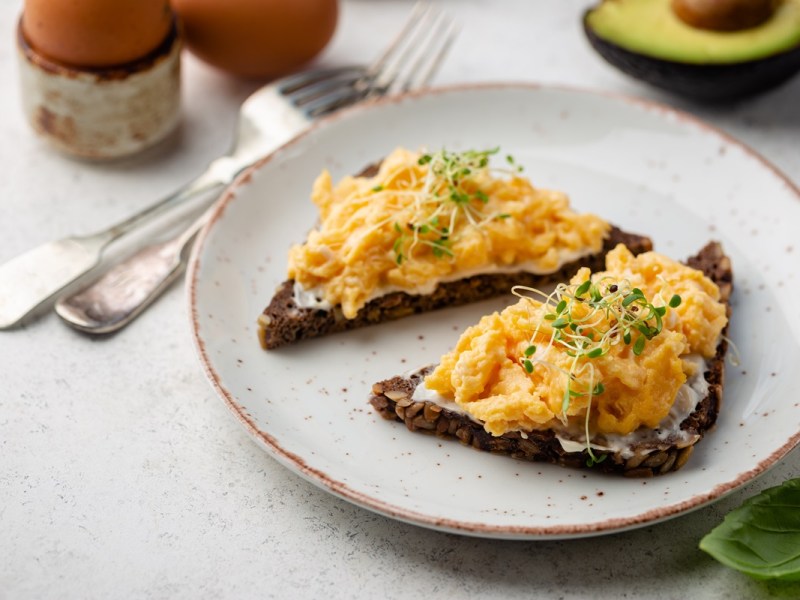 Zwei Brotscheiben mit Rührei mit Quark auf einem hellen Teller. Das Ei ist mit Sprossen garniert.