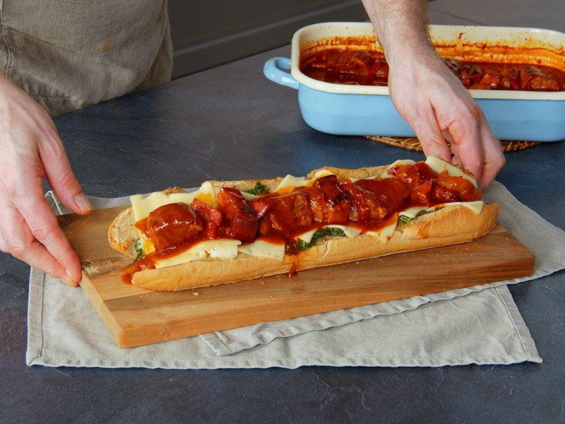 Ein Schaschlik-Sandwich auf einem Holzbrett. Unter dem Brett liegen Tischuntersetzer aus Stoff. Neben dem Schaschlik-Sandwich steht eine hellblaue Auflaufform mit Schaschlik-Soße und weiterem Schaschlik. Man kann zwei Hände erkennen, die nach dem Sandwich greifen.