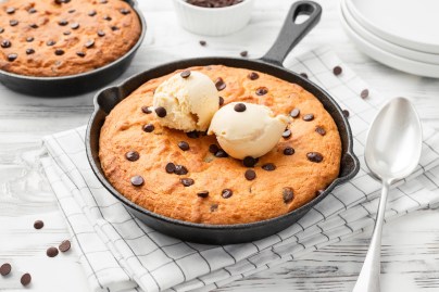 EIn Skillet Cookie, also Pfannenkeks, mit Schokotropfen in einer Pfanne, garniert mit zwei Eiskugeln. Daneben ein Esslöffel und eine weitere Pfanne mit einem Keks.