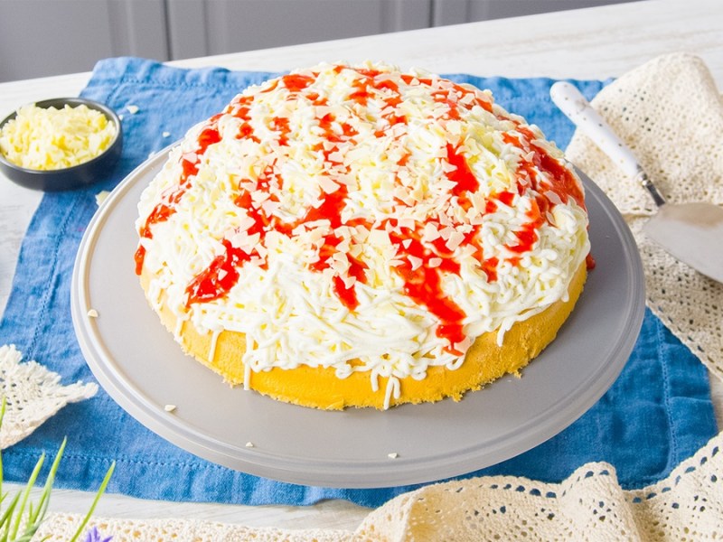 Ein Spaghettieis-Kuchen auf einem runden grauen Untersetzer. Er steht auf einem blauen Küchentuch. Drumherum verteilt liegt ein weitere Tuch im Hintergrund stehen Raspel von weißer Schokolade.