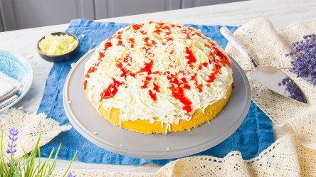 Ein Spaghettieis-Kuchen auf einem runden grauen Untersetzer. Er steht auf einem blauen Küchentuch. Drumherum verteilt liegt ein weitere Tuch im Hintergrund stehen Raspel von weißer Schokolade.