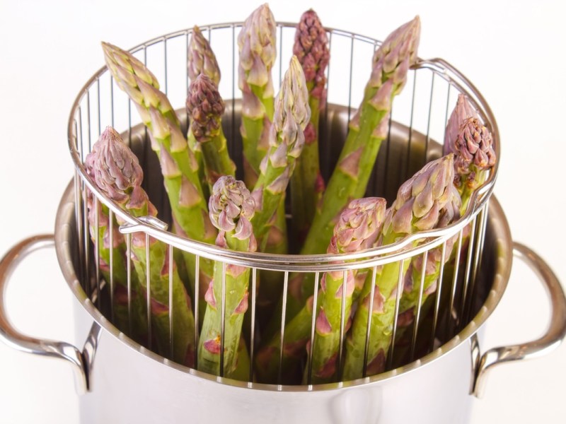 Ein Spargelkochtopf, aus dem der Garkorb etwas herausragt. Der Garkorb ist mit grünen Spargelstangen gefüllt.