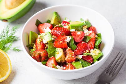 Eine Schüssel Spinatsalat mit Erdbeeren, Avocado und Feta. Daneben eine Gabel, eine halbe Zitrone und eine halbe Avocado. Nahaufnahme.