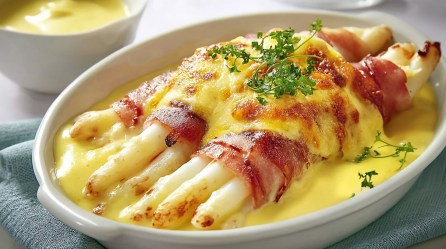 Überbackener Spargel mit Schinken und Sauce hollandaise in einer Auflaufform, bestreut mit Kräutern. Daneben eine Schale mit der Sauce hollandaise. Nahaufnahme.