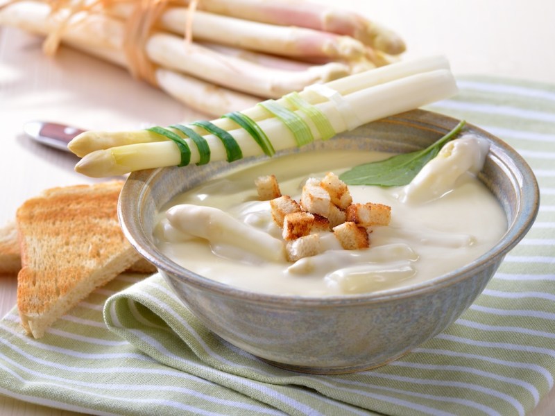 vegane Spargelcremesuppe in einer Schüssel, garniert mit Spargelspitzen und Crôutons. Daneben Toastbrotscheiben und Spargelstangen.