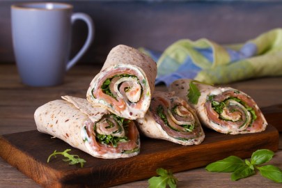4 Wraps mit Lachs und Meerrettich auf einem Holzbrett, dahinter eine Tasse und ein Geschirrtuch.