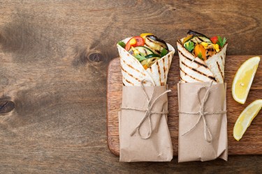 2 Wraps vom Grill gefüllt mit Gemüse und Koriander, eingewickelt in Backpapier, auf einem Brett, daneben zwei Zitronenspalten.