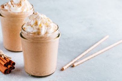 Zwei Gläser Zimtschnecken-Milchshake mit Sahne, daneben Zimtstangen und zwei Strohhalme.