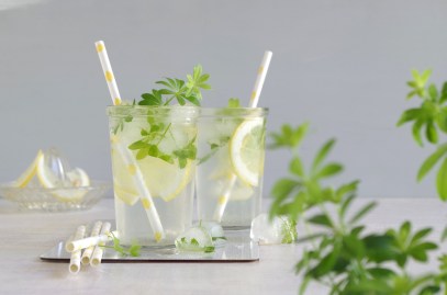 Zwei Gläser mit alkoholfreier Maibowle stehen auf einem weißen Tisch. Vor ihnen liegen ein paar Eiswürfel. Im Vordergrund ist noch frischer Waldmeister.