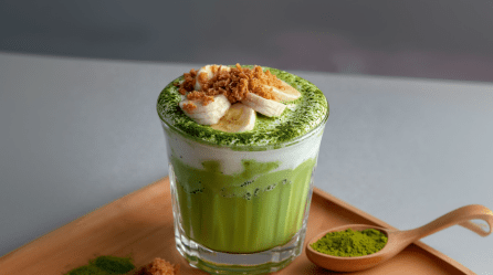 Ein Glas mit Banana-Pudding-Matcha auf einem Holztablett. Rechts neben dem Glas liegt ein Holzlöffel mit Matchapulver. Links liegt ebenfalls etwas Matchapulver sowie Keksbrösel