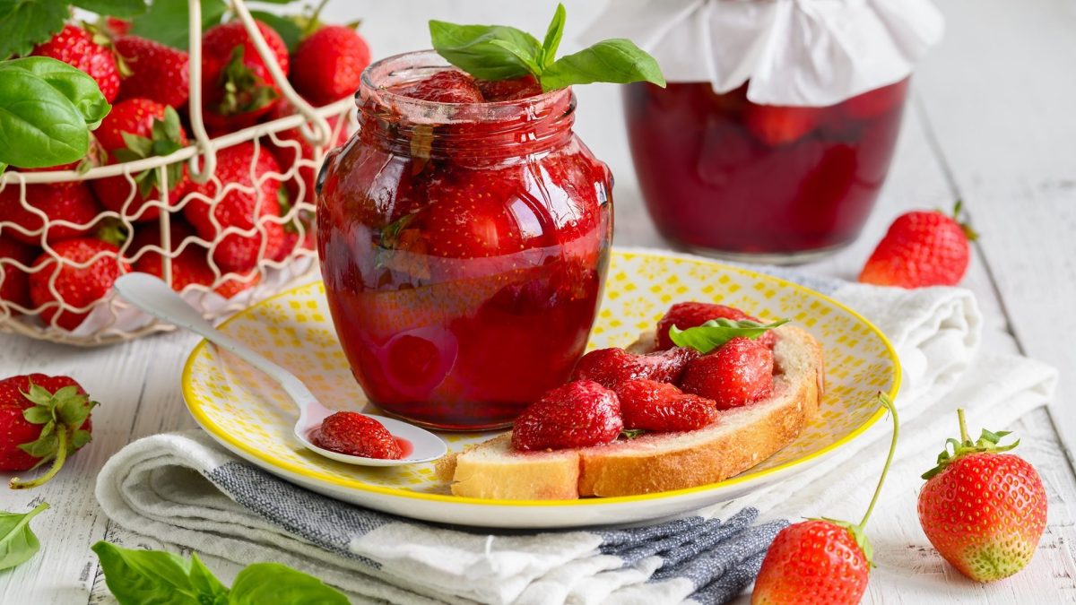 Ein offenes Glas Erdbeer-Basilikum-Marmelade mit Erdbeeren und Basilikumblättern drum herum.